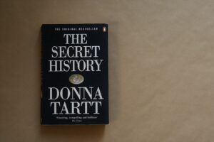 Das Buch "The Secret History" mit schwarzem Cover mit viel weißem Text von Donna Tartt liegt auf beigem Hintergrund.