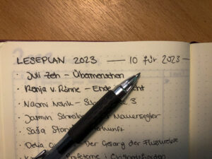 Ein Notizbuch liegt auf einem Holztisch. Darin steht eine Liste mit Büchern.